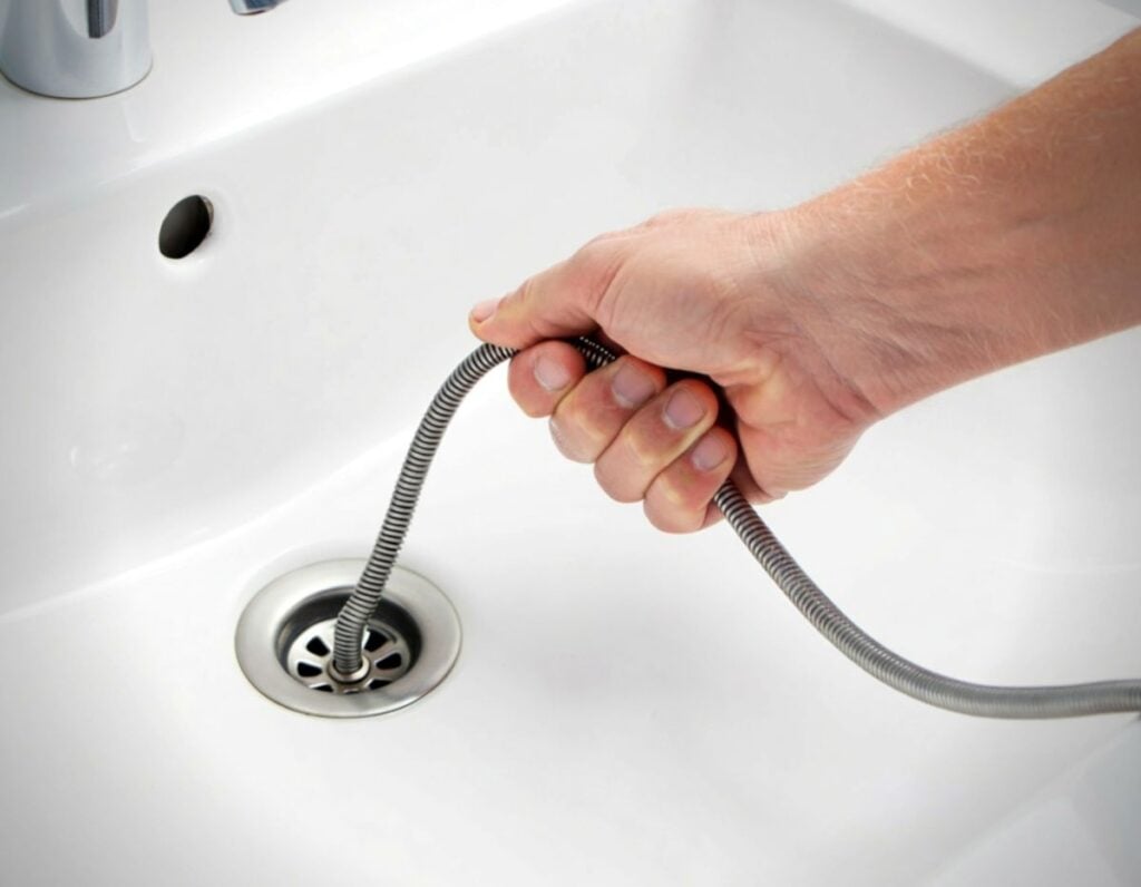 A hand uses a plumbing snake to clear a clogged drain in a white sink