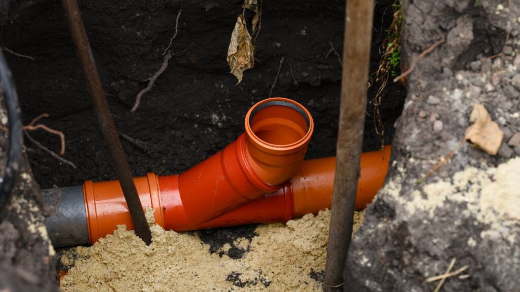 An orange plastic sewer pipe fitting installed in a deep dirt trench
