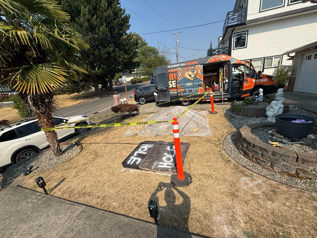 Brewer Sewer van beside a front yard sewer repair site, with caution tape, cones, and boards covering the dig.